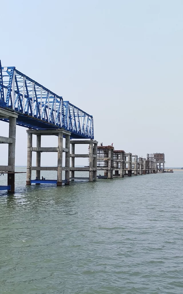 Blue steel truss access bridge on concrete piers extending over water as part of the Purulia piped water supply project in India.