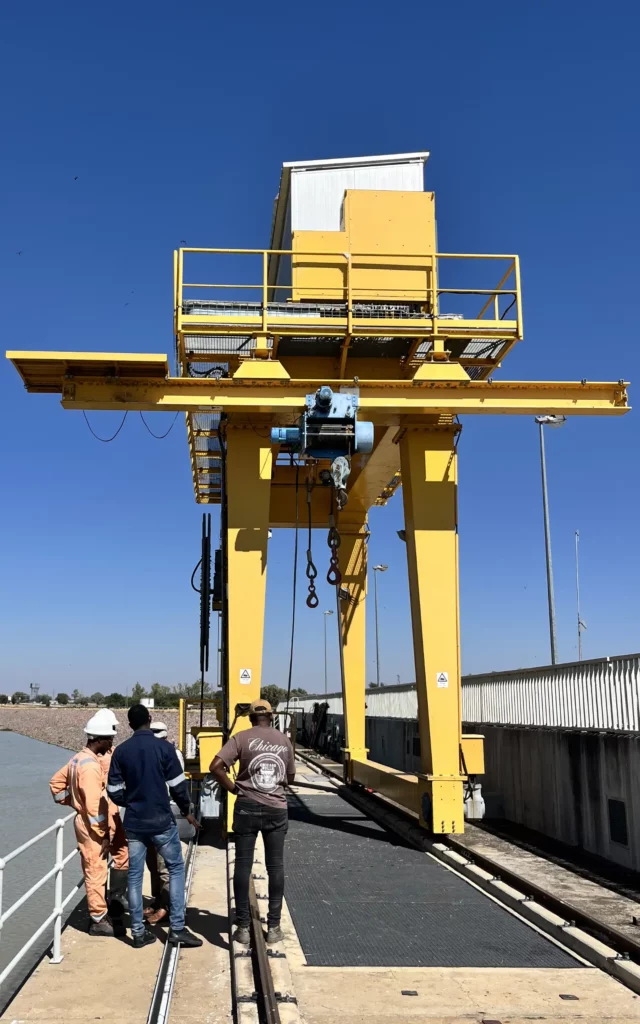 Rail-mounted yellow gantry crane and hoist above a dam intake platform, with three workers inspecting the equipment Rail-mounted yellow gantry crane and hoist above a dam intake platform, with three workers inspecting the equipment