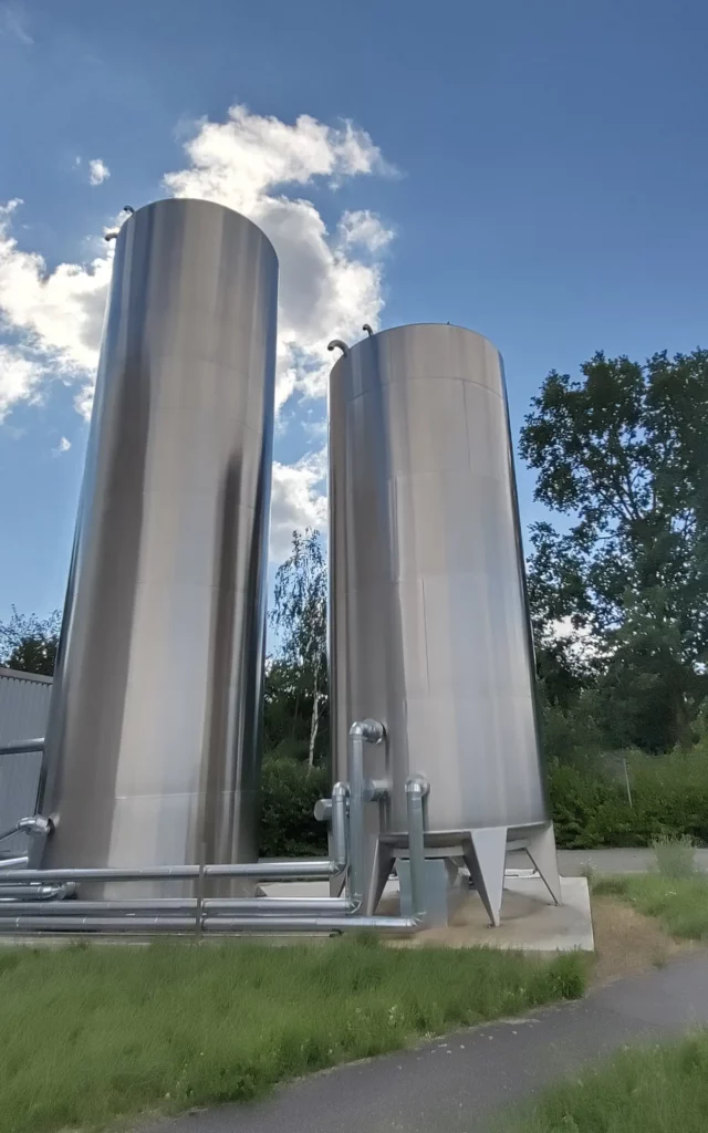 Vertical stainless-steel tanks connected by external pipework beside an industrial building. Vertical stainless-steel tanks connected by external pipework beside an industrial building.